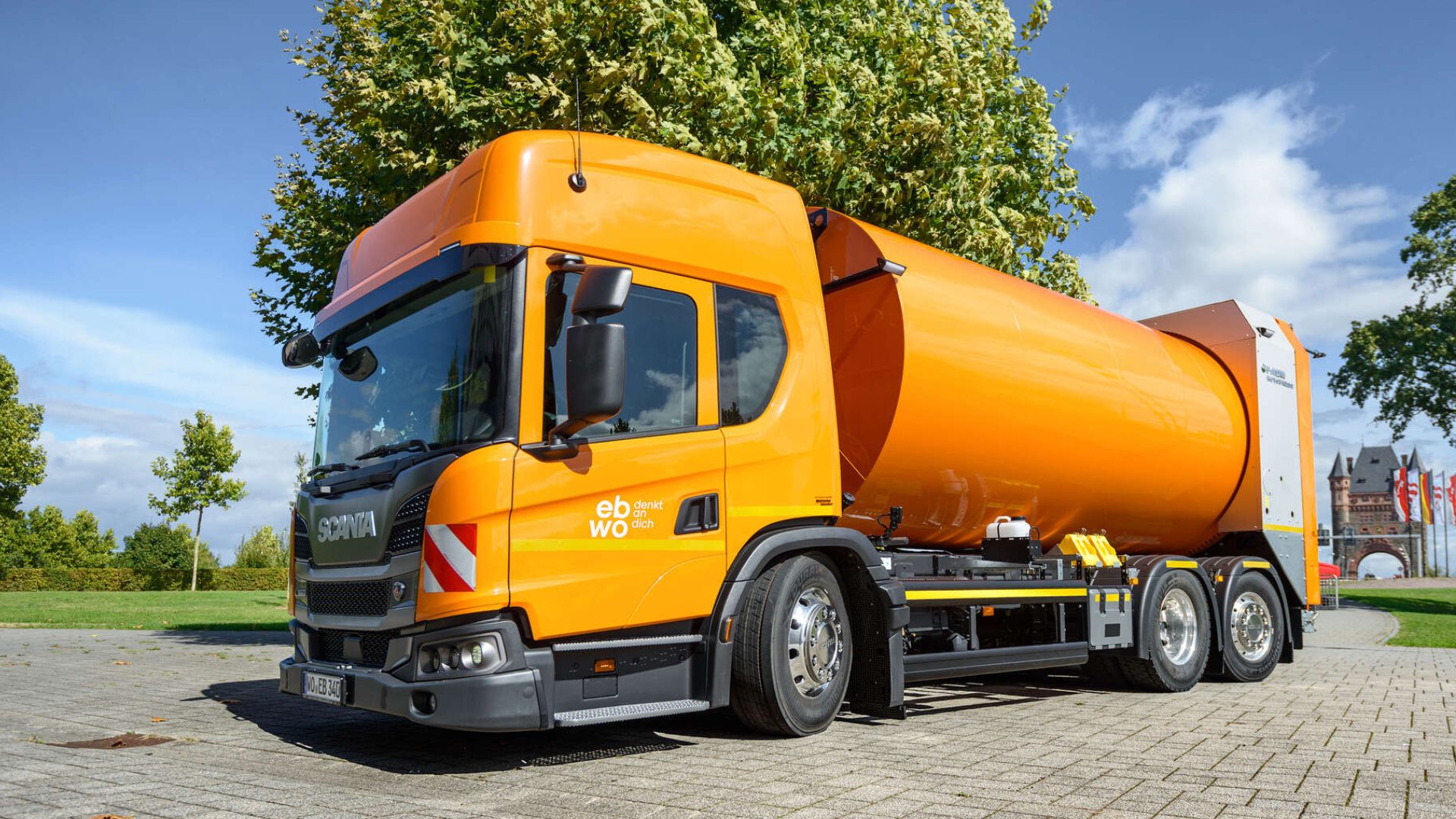 Neues Müllfahrzeug der ebwo AöR: ein Niederflurmodell mit Drehtrommelaufbau. Das Foto zeigt das orangefarbene Fahrzeug auf dem Küchlerplatz, im Hintergrund sieht man den Turm der Rheinbrücke.